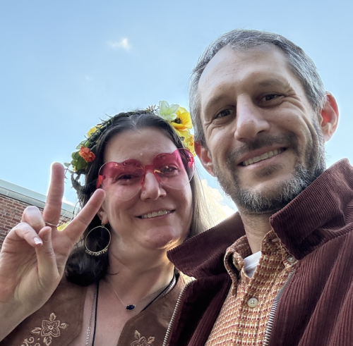 Aaron Podolner and a friend smiling outdoors for a selfie, wearing a flower crown and heart-shaped pink glasses