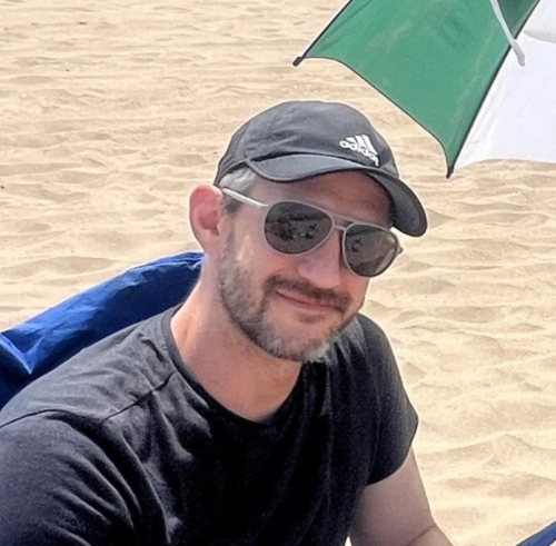 Aaron Podolner sitting on a beach under an umbrella, wearing a black t-shirt, cap and sunglasses