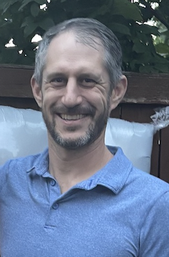 Aaron Podolner smiling, with a short grey beard and blue polo shirt, foliage in the background
