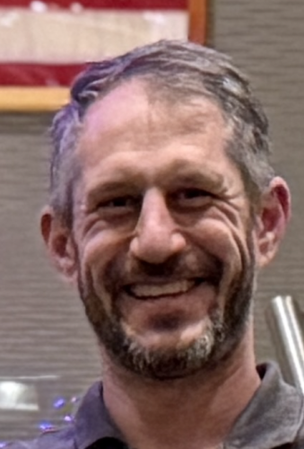 Professional headshot of Aaron Podolner with short grey hair, smiling broadly against a solid blue background