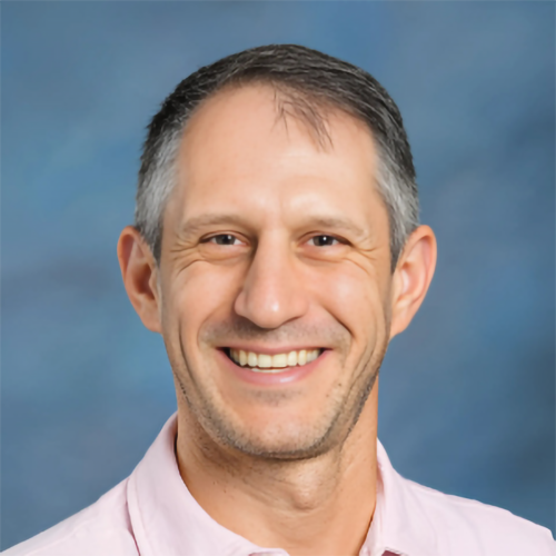 Aaron Podolner in an orange and blue striped shirt, speaking in a science classroom