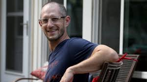 Aaron Podolner wearing glasses, sitting in a chair outdoors and smiling at the camera