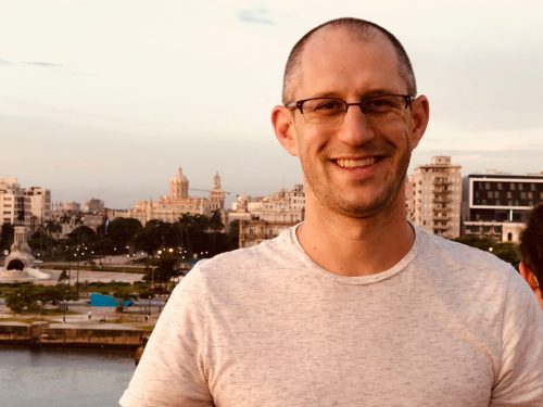 Aaron Podolner with glasses smiling in the foreground, with a city skyline and water behind him at dusk